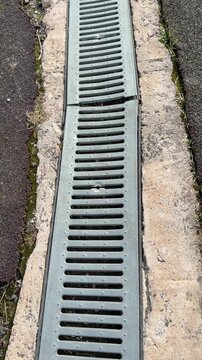 Galvanized drainage grate along stadium curb, long metal channel between asphalt and concrete, repeating slot pattern, leading