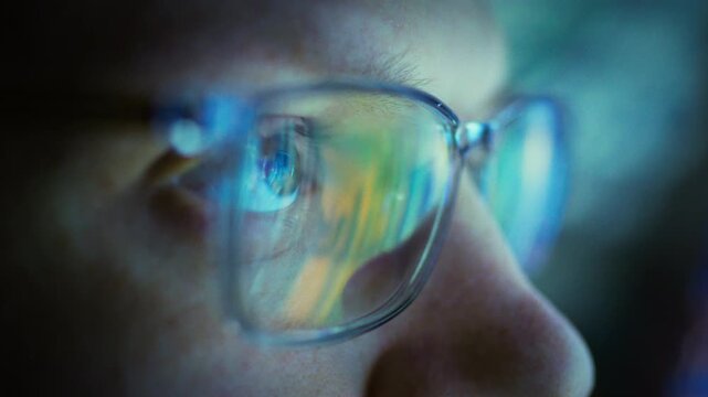 A programmer works through the night, neon-lit glasses bathing his face in electric glow as he codes. The software worker's focused stare mirrors the vibrant, after-hours tech atmosphere