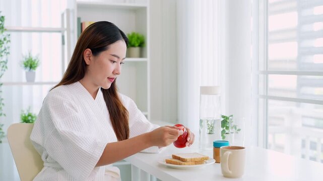 Young woman in bathrobe preparing breakfast putting jam on bread .