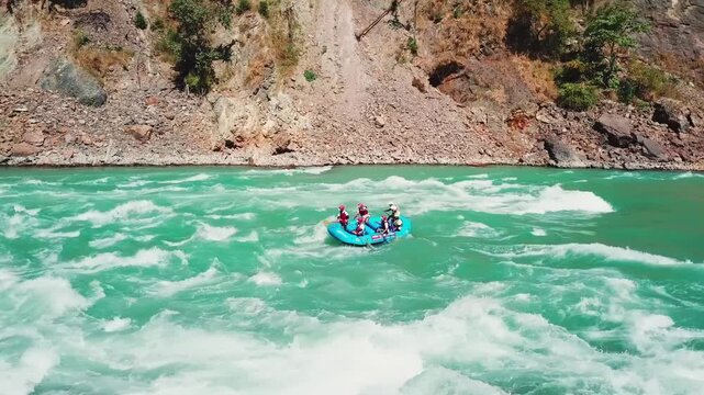 Rafting team paddling through turquoise rapids on the Ganges River in a mountain canyon near Rishikesh. Smooth forward tracking aerial shot follows the raft downstream past rocky terrain and forested.