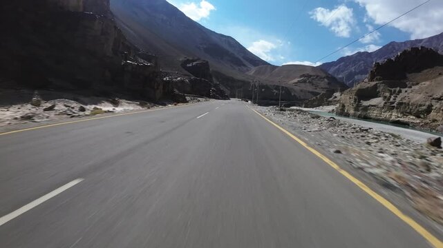 Ladakh's high-altitude highway cuts through arid mountain terrain alongside a glacial river. Low-altitude FPV drone tracking shot moves forward along the centerline with ascending perspective.