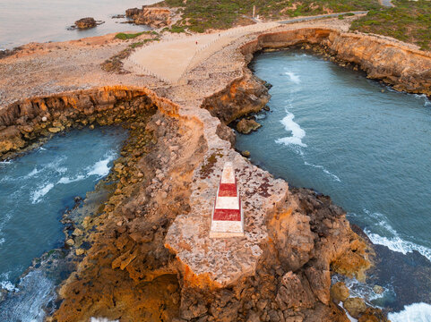 Aerial view of an obelisk monument high on a eroded cliff top surrounded by the sea