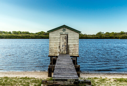 Old weathered wooden boat house with crooked jetty on river with blue sky and green vegetation