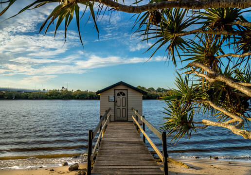Timber boat shed and jetty on sandy river shoreline with pandanus tree, blue sky and clouds