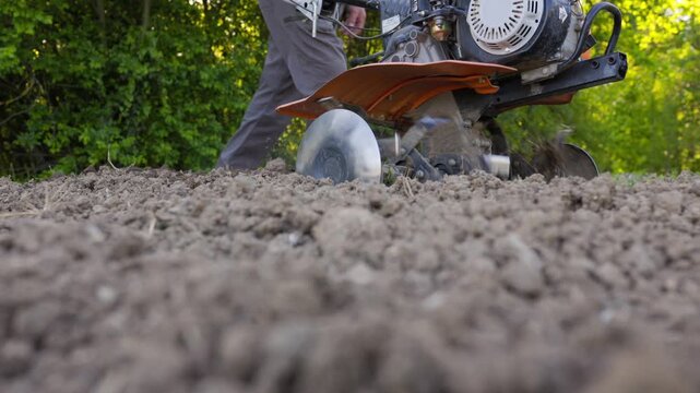 A ground-level shot focusing on the soil being turned and loosened by a gasoline-powered cultivator, with the machine and operator moving through the frame.