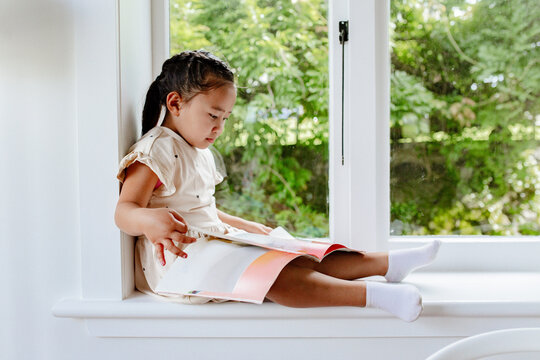 Young samoan girl reading a book while sitting by the windowsill