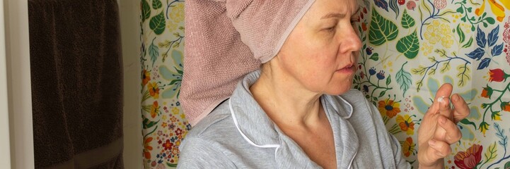 A 50-year-old woman prepares her skin by applying cream in her bathroom. She wears a towel on her head and stands in front of a colorful shower curtain during her morning routine, banner © pundapanda