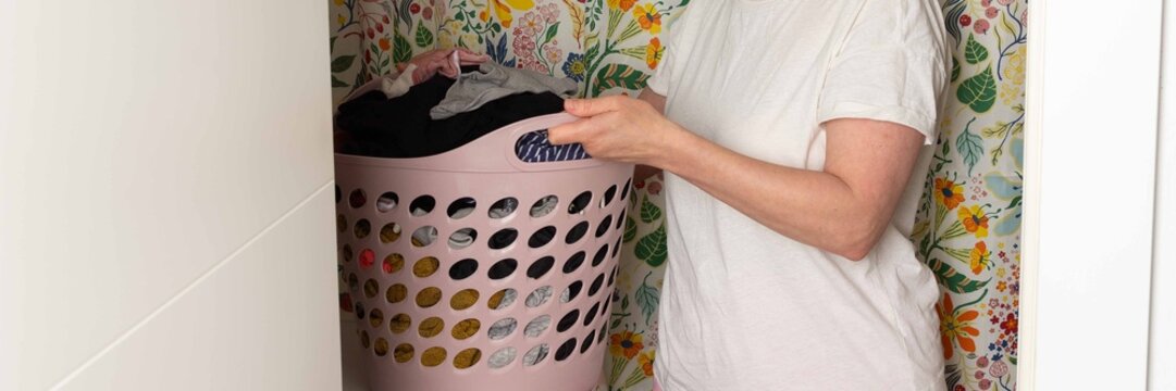 A woman removes clean laundry from the washing machine in a laundry room. She holds a basket filled with clothes and looks down at the laundry while standing on tiled floor, banner