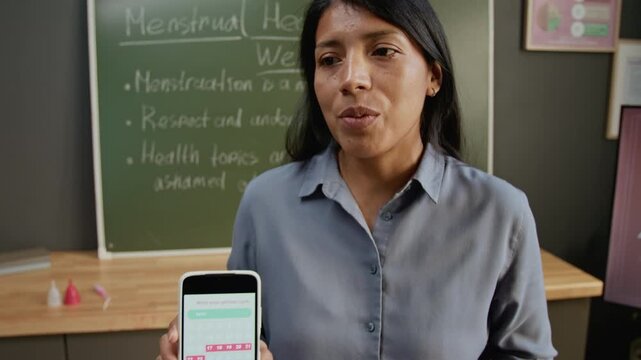Medium close-up of hands and face of cheerful Hispanic female teacher demonstrating smartphone with menstruation cycle tracking app while educating schoolgirls about reproductive health care