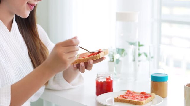 Young woman in bathrobe preparing breakfast putting jam on bread .