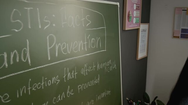 Medium close-up of professional Hispanic female teacher drawing on chalkboard while introducing discussion on STIs at reproductive health course, promoting awareness and responsibility among students
