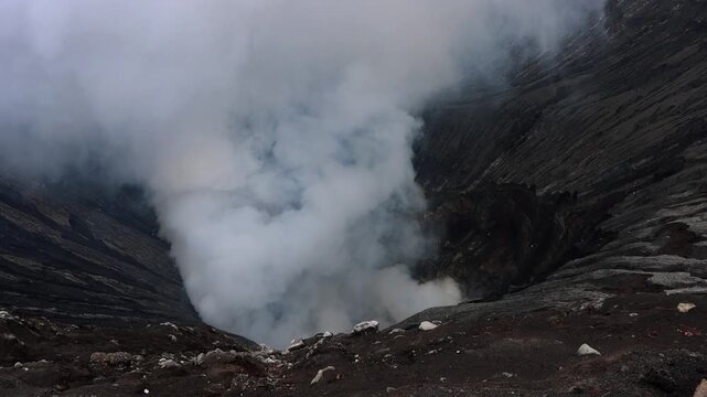 Inside Mount Bromo volcano crater with dense volcanic steam rising from active vents. Dark ash-covered crater walls and rugged volcanic terrain in East Java, Indonesia.