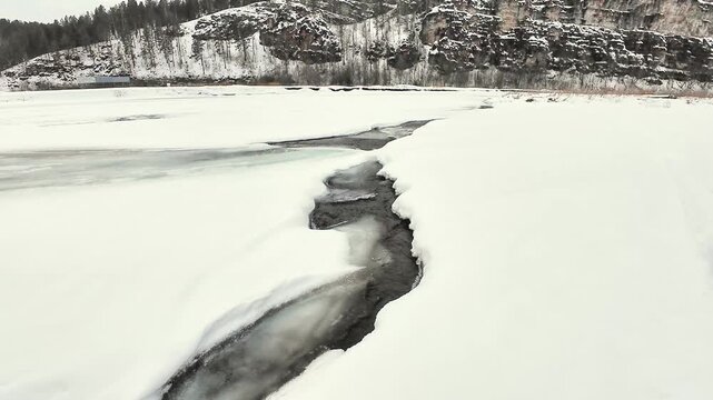 Frozen River Crack Cutting Through Broad Snowfield, Wide Aerial Sweep Revealing Dark Fissure And Thin Ice, Distant Rocky Bluff And Tree Line On Horizon, Subtle Underice Current And Textured