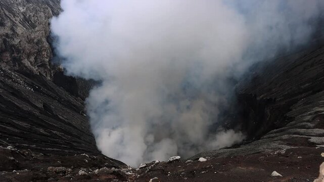 Inside Mount Bromo volcano crater with dense volcanic steam rising from active vents. Dark ash-covered crater walls and rugged volcanic terrain surround a dramatic geothermal landscape in East Java