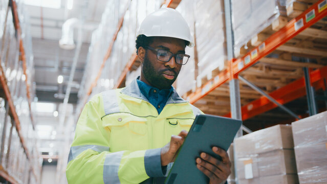 Portrait African American technician wearing glasses tapping on tablet while standing near stacked pallets. Reviewing digital checklist or checking inventory database in modern warehouse.