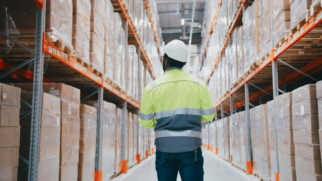 Warehouse technician in high-visibility gear walking away while holding tablet. Surrounded by stacked packages on both sides. Navigating aisles and updating inventory records during inspection.