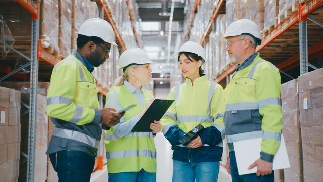 Logistics team wearing high-visibility jackets and helmets holding devices and clipboards while planning in warehouse. Four professionals standing surrounded by pallets and industrial racks.