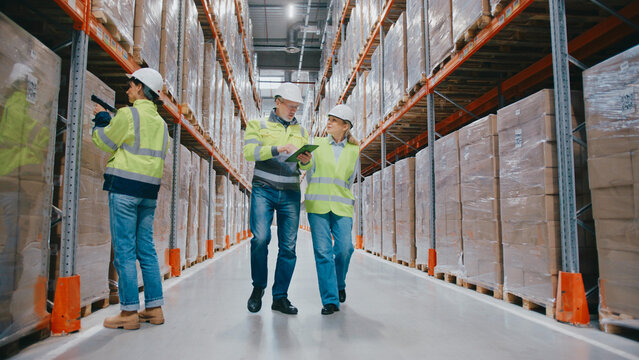 Warehouse workers wearing safety gear operating in large distribution center. One scanning boxes, while two colleagues inspect shelves and discuss storage with clipboard and hand gestures.