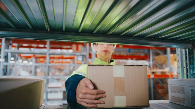 Female warehouse worker in safety gear holding box and barcode scanner while looking toward shelf. Standing in front of industrial racking system filled with labeled packages. Commercial center.
