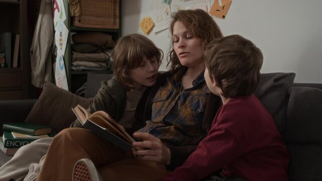 Low angle shot of caring mother reading book for her children, sitting together on couch in poor cluttered apartment