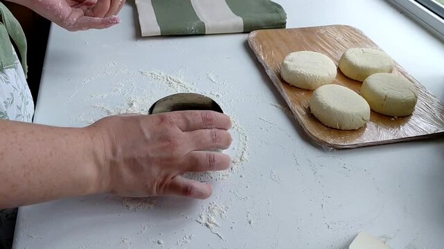 Hands of a female chef preparing homemade syrniki (cottage cheese pancakes) by coating them in flour on a wooden board before frying. Home cooking, traditional recipes, and culinary preparation.