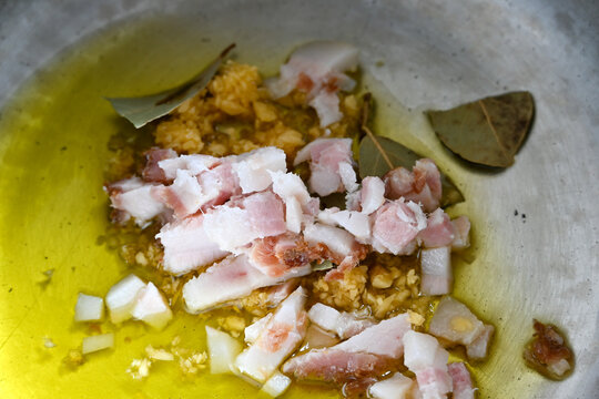 Close-up of chopped pancetta, minced garlic, and aromatic bay leaves being saut&eacute;ed in extra virgin olive oil inside a rustic metal pot, preparing a flavorful base for a traditional Italian meal
