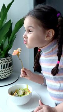 A little girl eats pasta with vegetables near the window. Funny child eats vegetables and laughs