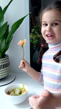 A little girl eats pasta with vegetables near the window. Funny child eats vegetables and laughs