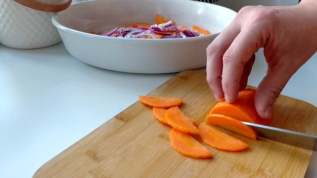 A person slices vibrant orange carrots with a knife on a rustic wooden cutting board