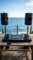 Fototapeta premium Professional DJ equipment setup on a wooden pier overlooking a blurred tropical blue ocean under a clear summer sky.