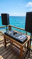 Fototapeta premium Professional DJ equipment setup on a wooden pier overlooking a blurred tropical blue ocean under a clear summer sky.