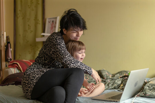 mother and child on bed browsing laptop, relaxed bedside scene with affectionate cuddle, mother multitasking as freelance creator while child watches, soft pillows and blanket, warm lamp