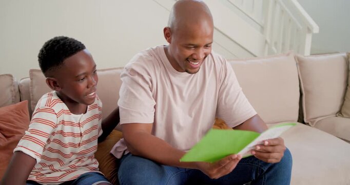 African American dad-son hugging on sofa as child placing hand, dad reading green card, sipping mug