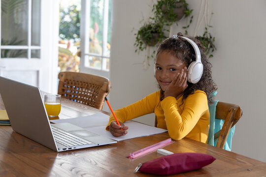 Schoolgirl sitting at dining table writing with orange pencil near laptop wearing white headphones