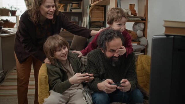 Father and his older son playing video games, mother resting hands on their shoulders, while younger son playfully covering his fathers eye with his hand in small crampred apartment
