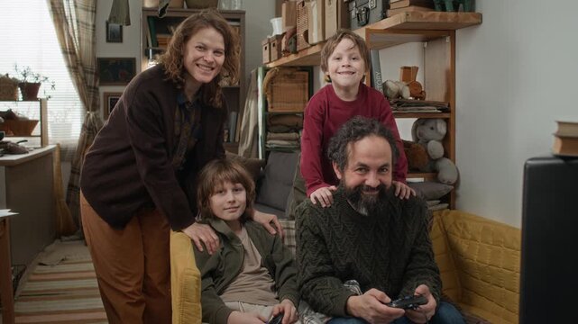 Portrait shot of mom, dad, and their two sons smiling and looking at camera while sitting on couch with video game controllers in poor apartment