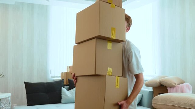 Close up of handsome Caucasian red-haired guy holding many cardboard boxes in hands. Boxes falling on the floor. Moving out the apartment. Relocation concept. Real time, indoors