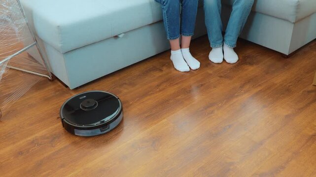 Modern vacuum robot cleaning at home. Close up. Cleaner vacuuming parquet in room. Couple raising their legs while sitting on couch. High angle view. Cleaning robot. Technology concept. Real time