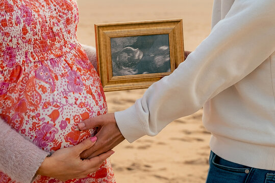 Pregnant woman  with husband on sand near ocean. Mother and father hugging in anticipation of baby and holding photo of baby's girth in golden wooden frame. Pregnancy planning in sunny Portugal