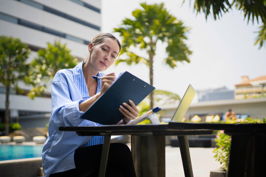 A young entrepreneur woman is working on her computer and document in by the resort pool in summer time. Remote working anywhere, the concept of digital nomad. Top travel destination
