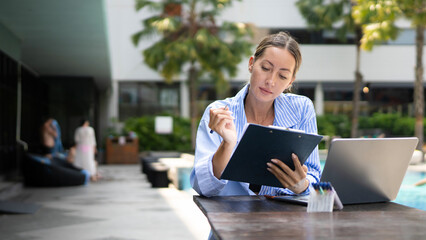 A young entrepreneur woman is working on her computer and document in by the resort pool in summer time. Remote working anywhere, the concept of digital nomad. Top travel destination © Stella 16