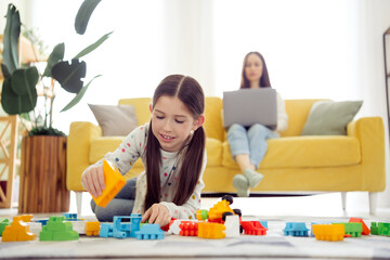 Obraz premium Girl playing with colorful building blocks on living room floor while mother works on laptop on yellow sofa — family, child play and remote work at home