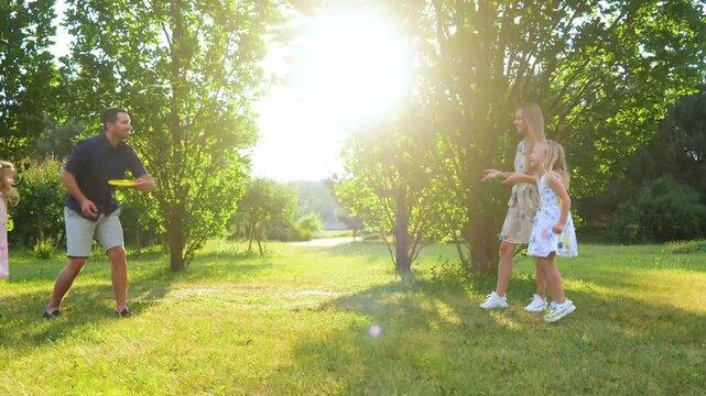 Caucasian family throwing frisbee disk in park outside. Happy girls jumping cheering for parents while mom and dad play with frisbee plate. Children and parents. Outdoor activity concept. Real time.