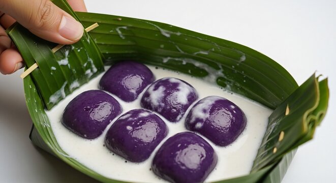 Hand opening a banana leaf wrap of Kue Bugis Mandi, soaked in thick white coconut sauce, plain white background.