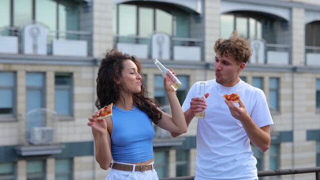 Happy friends biting pizza and holding bottles of beer, having fun at party on the rooftop in the city. Lifestyle, friendship, fun, open party concept. Real time