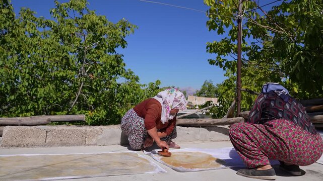 Two rural women spread grape pestil mixture onto cloth sheets on a sunny rooftop, capturing a traditional production stage, village life, manual food crafts, and authentic Anatolian culinary heritage.