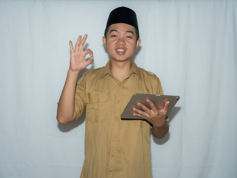 Young man wears traditional attire and cap. He smiles warmly while holding a tablet. His hand forms an "okay" gesture with confidence. Neutral background highlights his cheerful expression