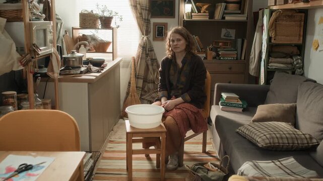 Portrait shot of woman sitting on chair in center of room crammed with household items, looking at camera with basin in front of her