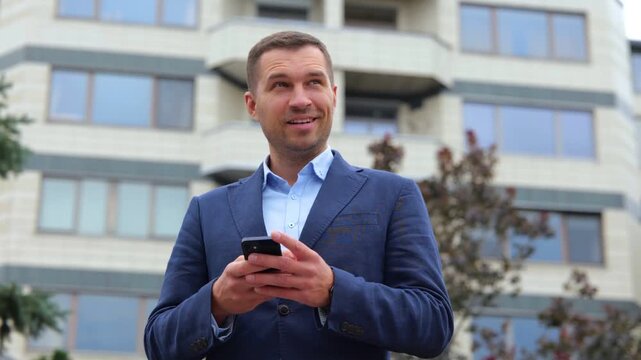 Happy handsome confident guy in suit typing on mobile phone and smiling in good mood. Close up of joyful businessman texting message on smartphone. Real time concept. Urban concept