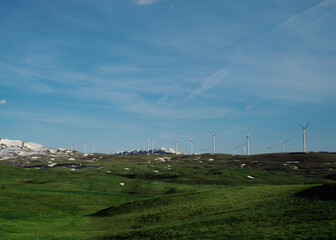 Fototapeta premium Wind turbines on green rolling hills with snow capped mountains in background, renewable energy and sustainable future concept